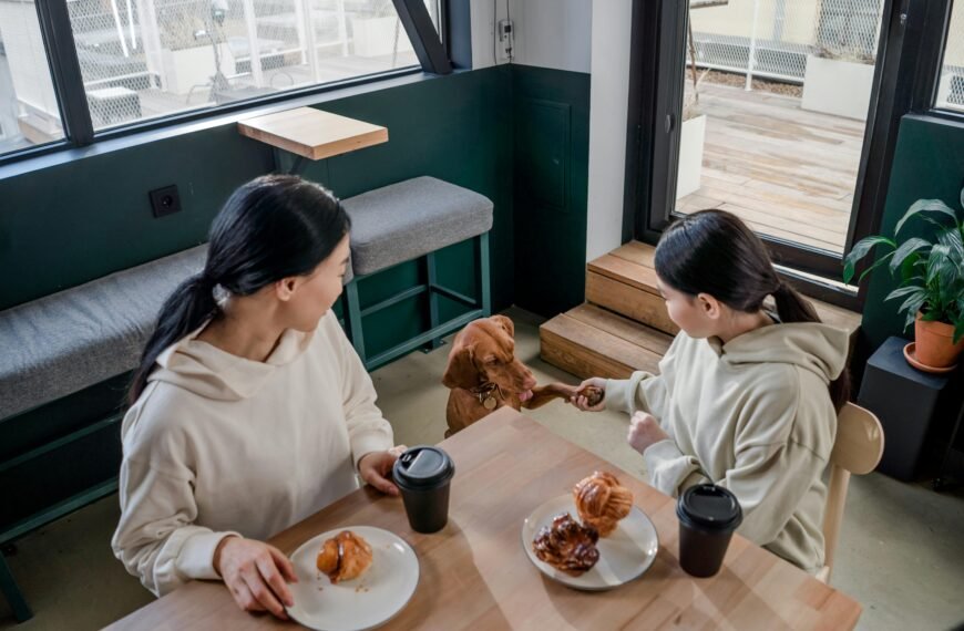 A mother and daughter share a warm moment with their dog in a café, enjoying coffee and pastries.